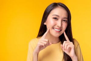 Woman in yellow shirt in front of yellow background smiling and pointing to her teeth